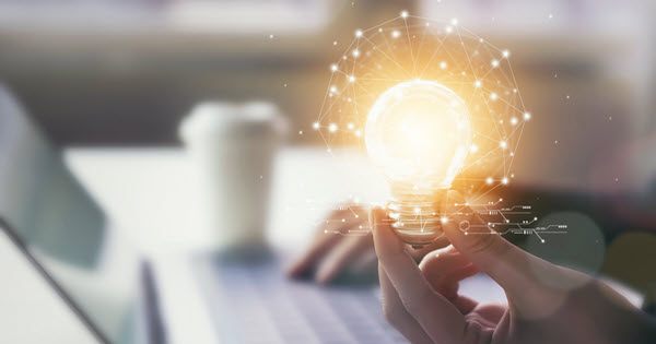 an online student working at a computer is holding a glowing lightbulb and there is a coffee cup in the background