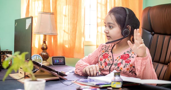 a young female student wearing headphones sitting at her desk at home taking an online class